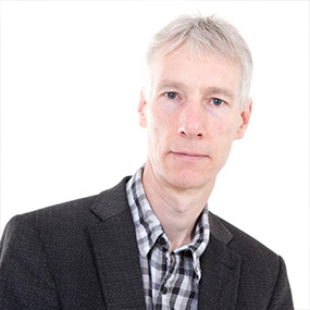 Man with short grey hair wearing a black and white checked shirt and dark jacket