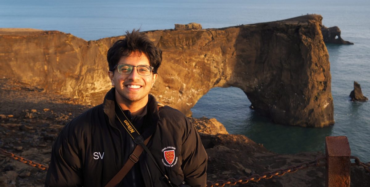 Sidharth wearing a black Oriel puffer jacket standing in front of a costal view.