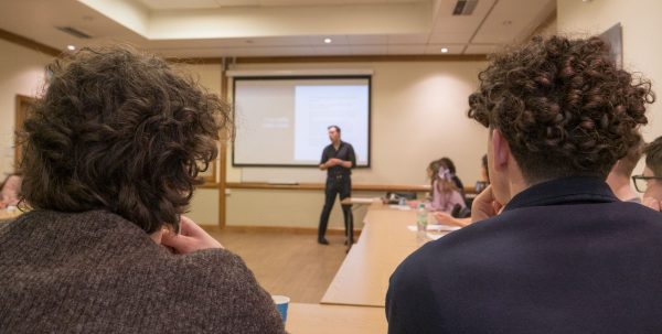 The photo is looking at a lecturer giving a talk in a classroom. in the photo is also the back of two students heads