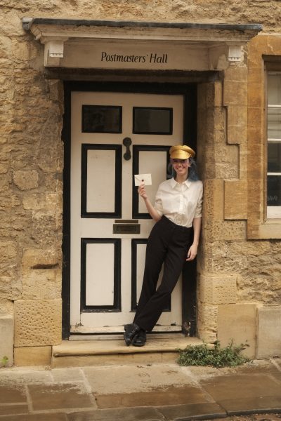 A young woman leaning against a doorway in a golden hat holding an envelope