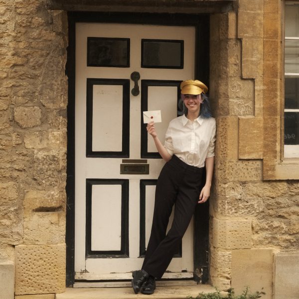 A young woman leaning against a doorway in a golden hat holding an envelope