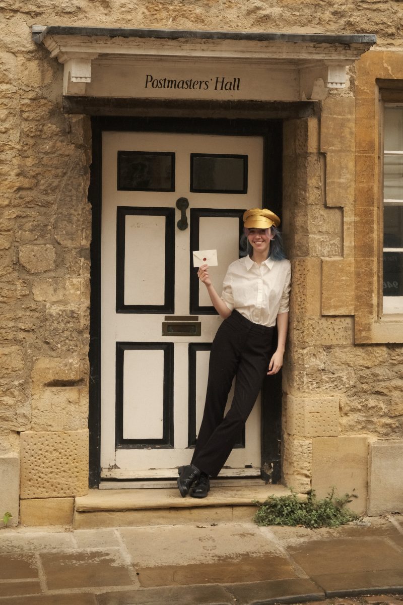 A young woman leaning against a doorway in a golden hat holding an envelope