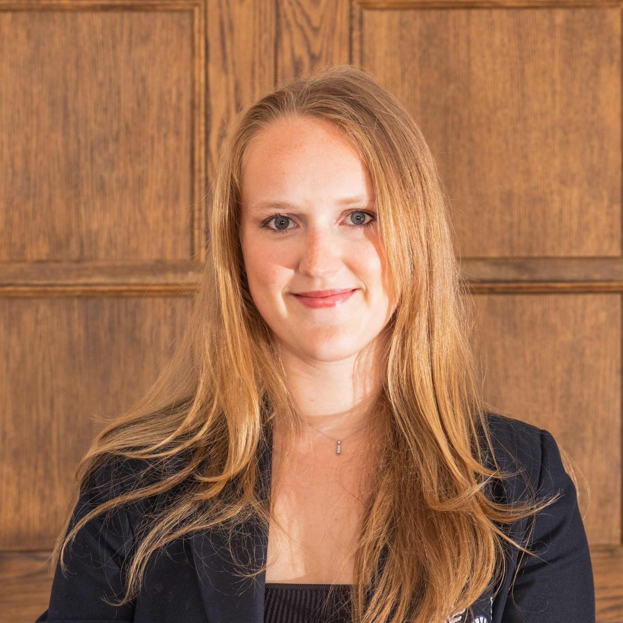 A woman with shoulder length blonde hair wearing a dark blue blazer standing in front of a wooden panelling background.