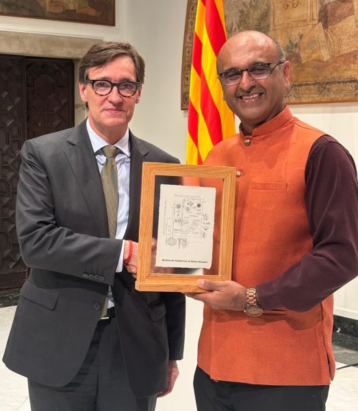 Two men standing and shaking hands and holding a certificate. The one on the left is wearing glasses and a suit and the one on the right is also wearing glasses and wearing a sleeveless orange Nehru jacket.