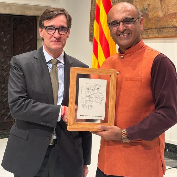 Two men standing and shaking hands and holding a certificate. The one on the left is wearing glasses and a suit and the one on the right is also wearing glasses and wearing a sleeveless orange Nehru jacket.